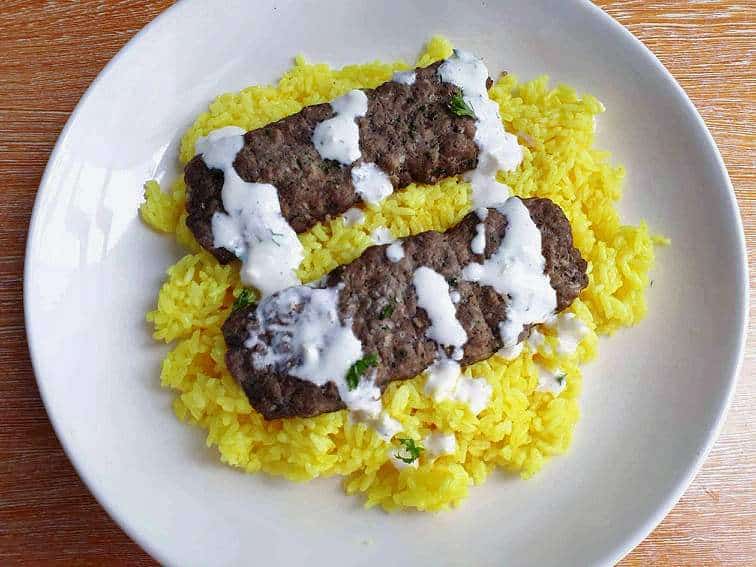 Jordanian Beef Kofta Kebab served with yellow rice, drizzle with white yogurt sauce and garnished with chopped parsley in a plate.