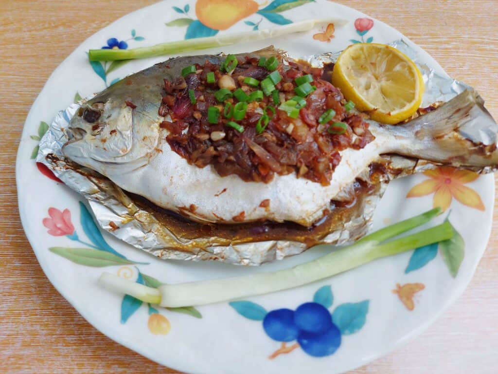 Whole baked pompano fish topped with onion-tomato soy sauce mixture with 2 onion leaves and 1 slice of lemon served on a plate