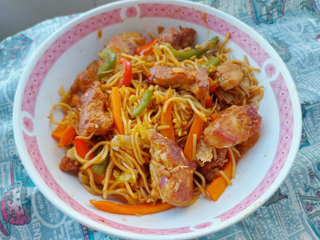 An indo-Chinese Chicken Manchurian noodle recipe mixed with fried boneless chicken breast, noodles,  julienned carrots and cabbages served on a plate