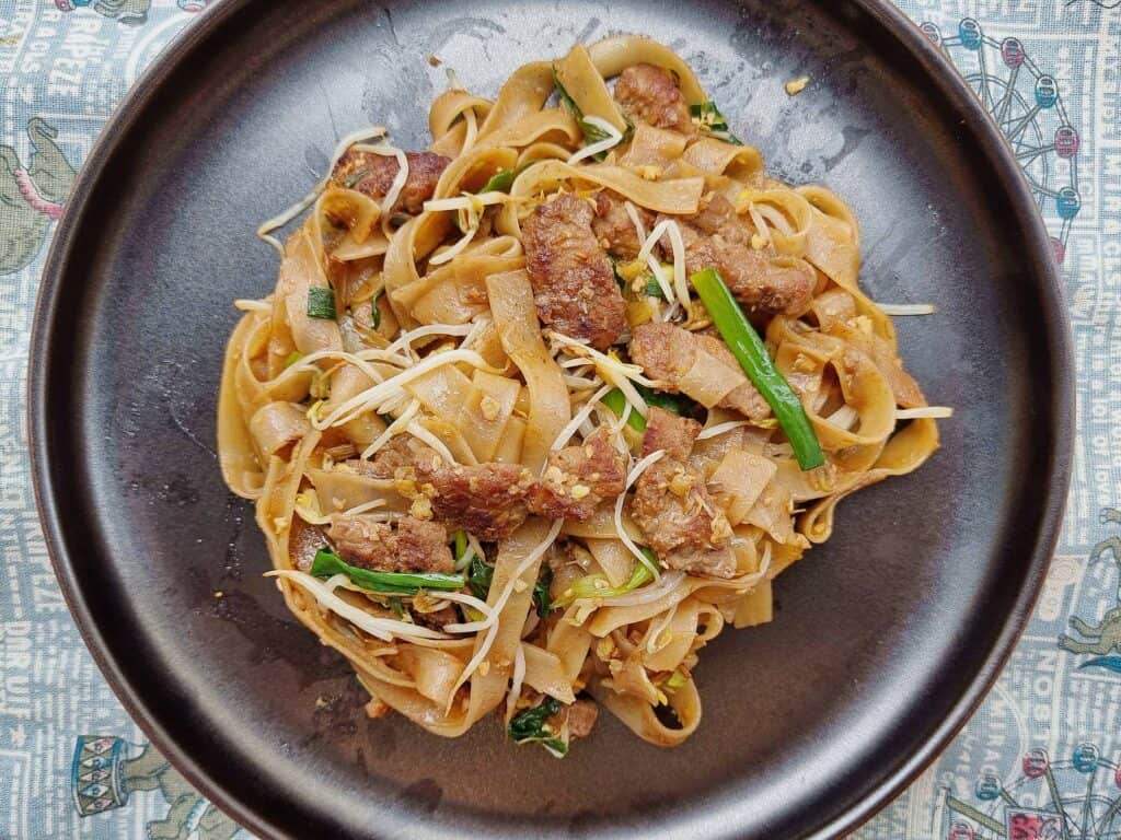 Singaporean stir fried noodle dish Beef Kway Teow mixed with thinly sliced beef, bean sprouts, green stalk, different sauces and rice noodles served on a plate.