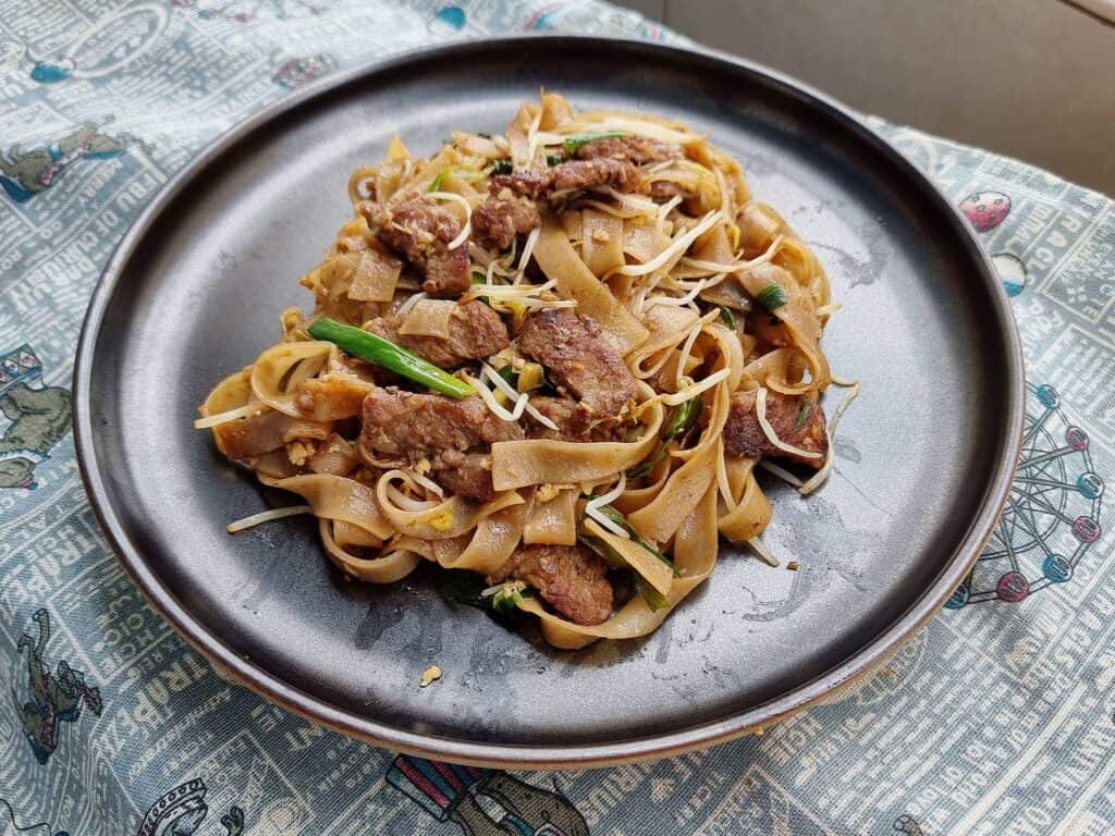 Singaporean stir fried noodle dish Beef Kway Teow mixed with thinly sliced beef, bean sprouts, green stalk, different sauces and rice noodles served on a plate.