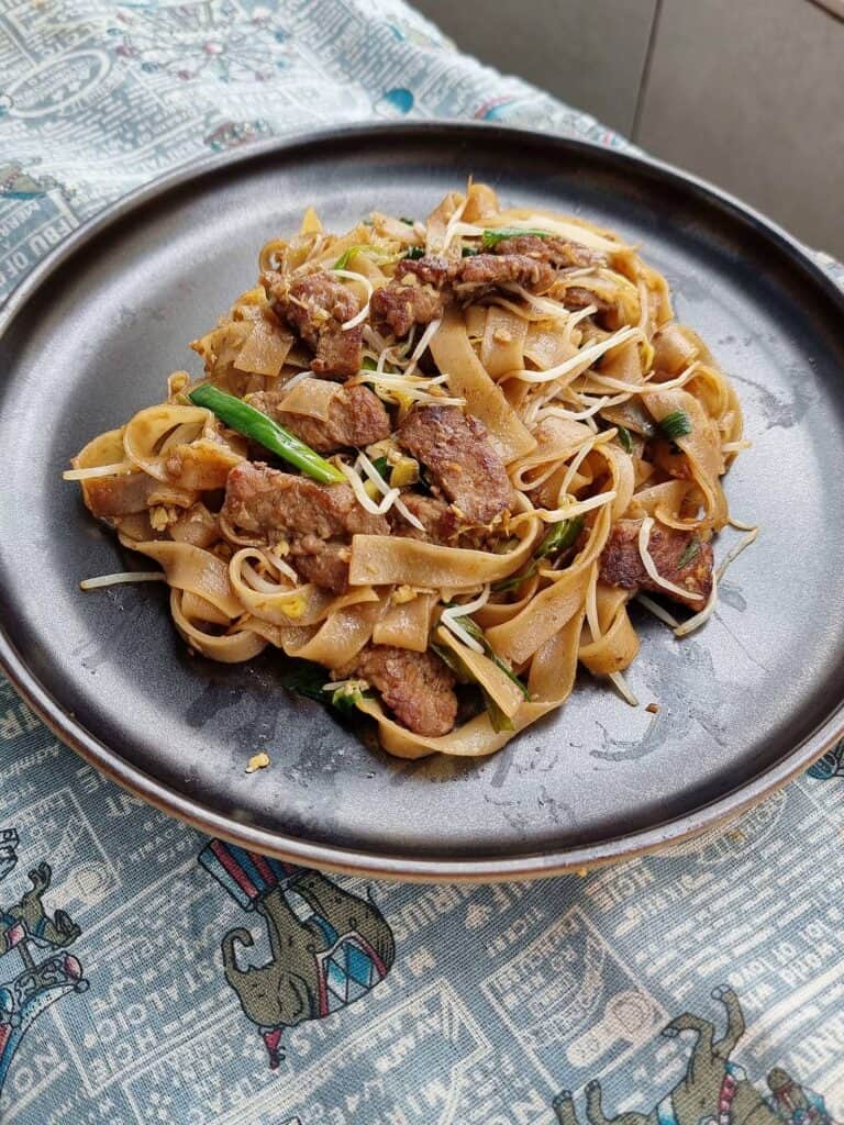 Singaporean stir fried noodle dish Beef Kway Teow mixed with thinly sliced beef, bean sprouts, green stalk, different sauces and rice noodles served on a plate.