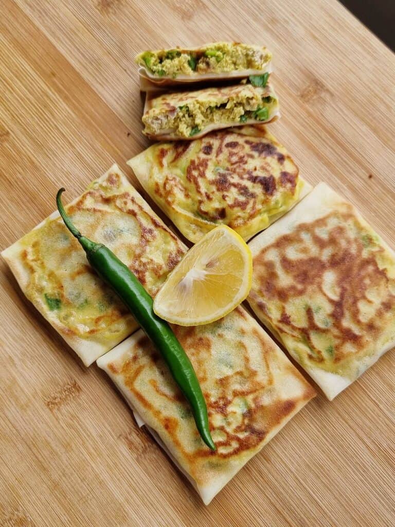 A golden Muttabaq pastry, layers of thin dough enveloping seasoned meat and greens, fried to crispy perfection in Saudi cuisine served with green chilies and lemon.