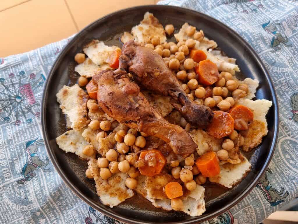 Chakhchoukha, a traditional Algerian dish, presents torn rougag flatbread soaked in a tomato-based broth with chicken, carrots, and chickpeas served on a plate