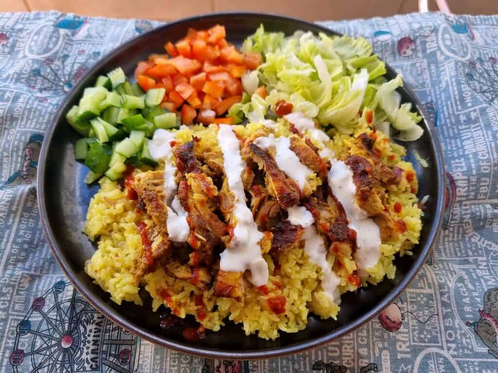 Shawarma Rice with baked marinated chicken, chopped tomatoes, carrots and lettuce and yellow rice topped with white sauce served on a plate.