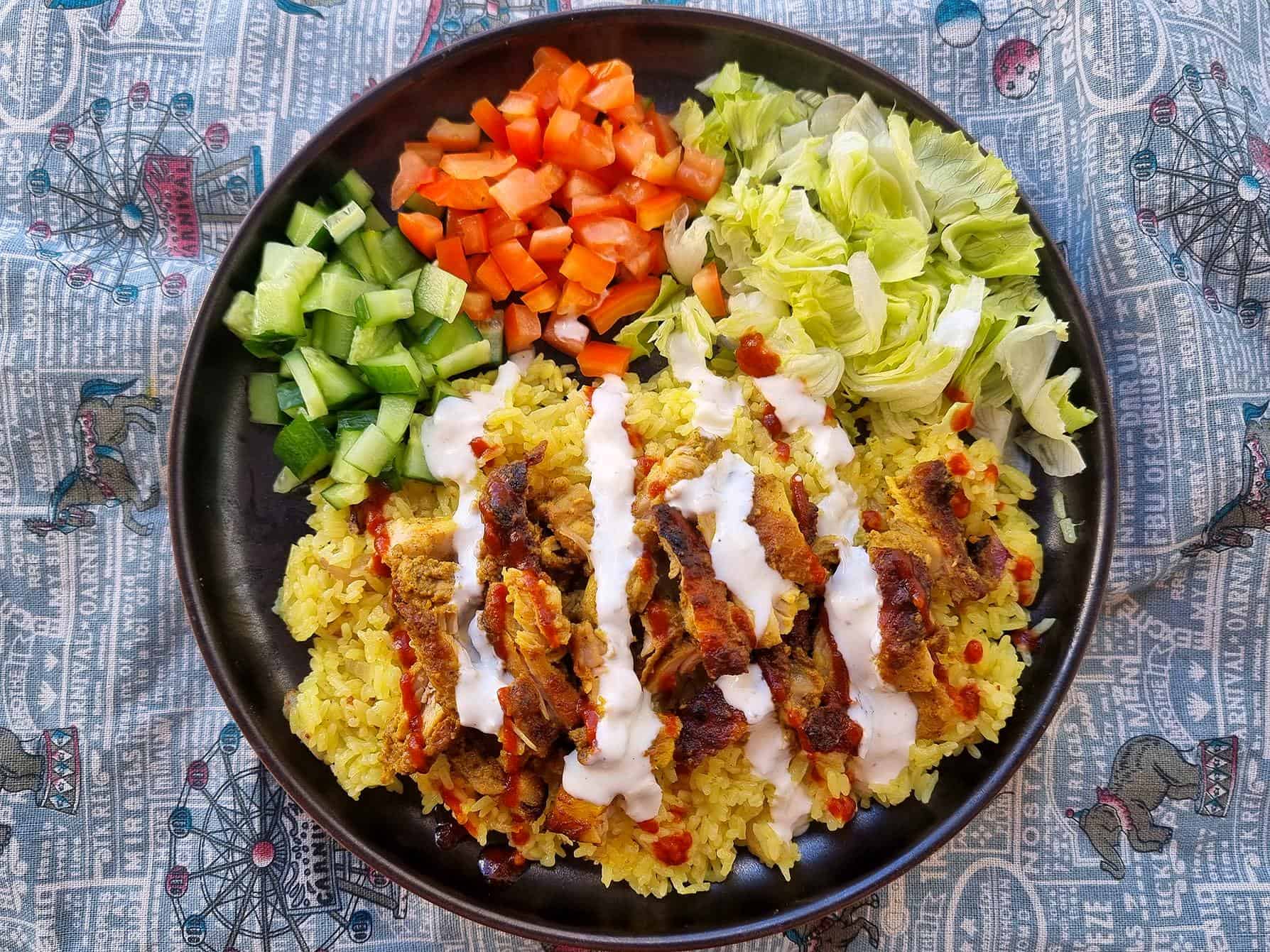 Shawarma Rice with baked marinated chicken, chopped tomatoes, carrots and lettuce and yellow rice topped with white sauce served on a plate.